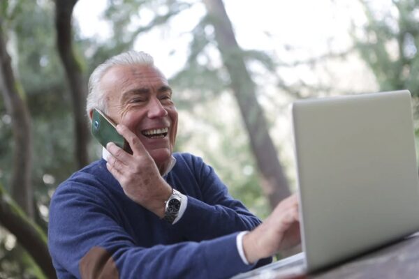 An elderly man working on his laptop while calling with his cell phone