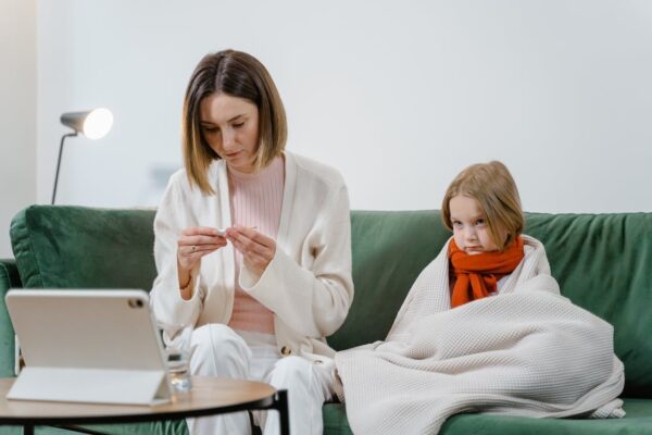 A woman reading the temperature on a thermometer with a sick child next to her on the sofa