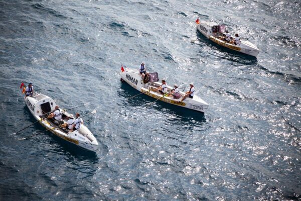 Three NoMan boats in open waters with people rowing.